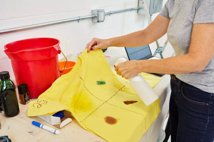 A hand is seen holding a spray bottle over a yellow t-shirt stained with a mark, placed next to a red bucket.