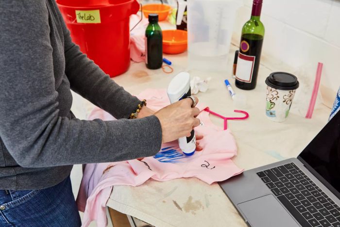 A hand applying Carbona Laundry Stain Scrubber to a stained t-shirt, placed next to a coffee cup and a bottle of wine.