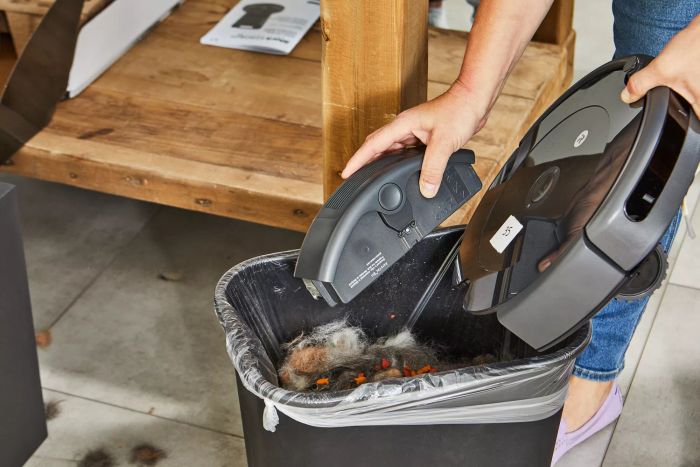 A person is seen holding a disassembled iRobot Roomba 694 Robot Vacuum, emptying its contents into a trash bin.