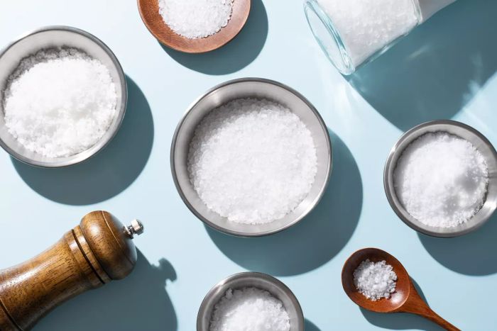 An assortment of sea salts, including fine, kosher, coarse, and flake salts, displayed in bowls and spoons against a soft blue background, viewed from above.