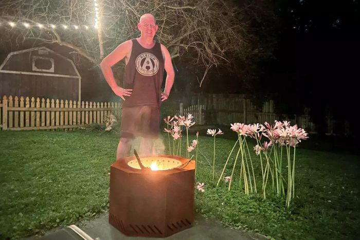 A man standing near the Titan Great Outdoors Corten Steel Dual-Flame Smokeless Fire Pit in an outdoor setting