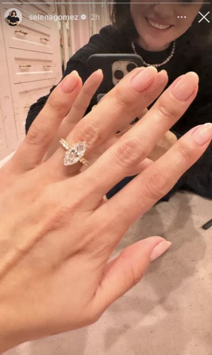 A mirror selfie captures a hand with manicured nails showcasing a sizable marquise-cut diamond ring, with only a portion of the face visible.