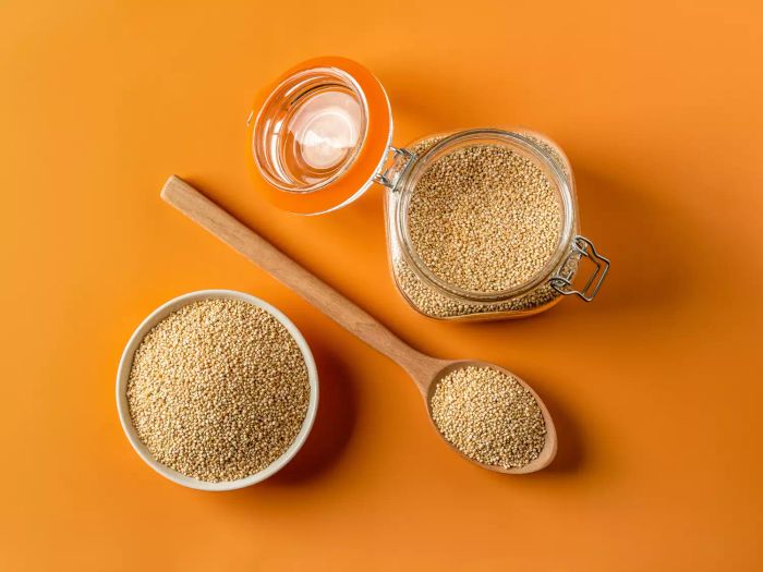 A close-up of raw quinoa in a bowl, accompanied by a wooden spoon and a glass jar, set against a vibrant orange backdrop. White quinoa seeds, ideal for a gluten-free diet, are showcased for cooking purposes.