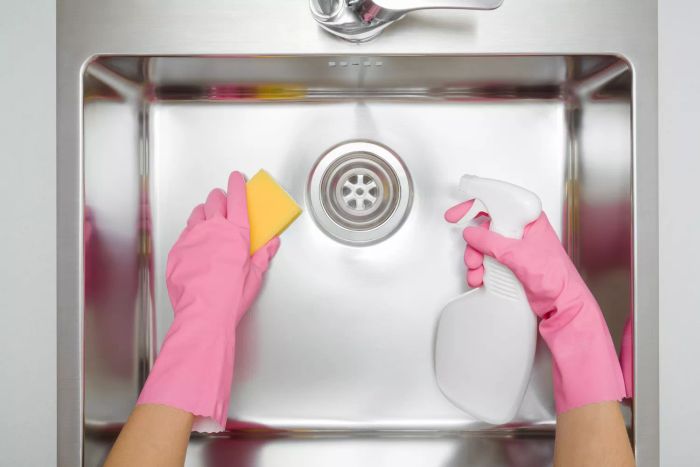 Close-up of a woman wearing pink rubber gloves, holding a white spray bottle and a yellow sponge while cleaning a light gray stainless steel kitchen sink. Top-down perspective.