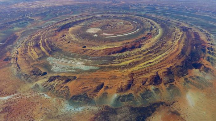An aerial perspective reveals a vast expanse of red, rugged mountains arranged in concentric circles.