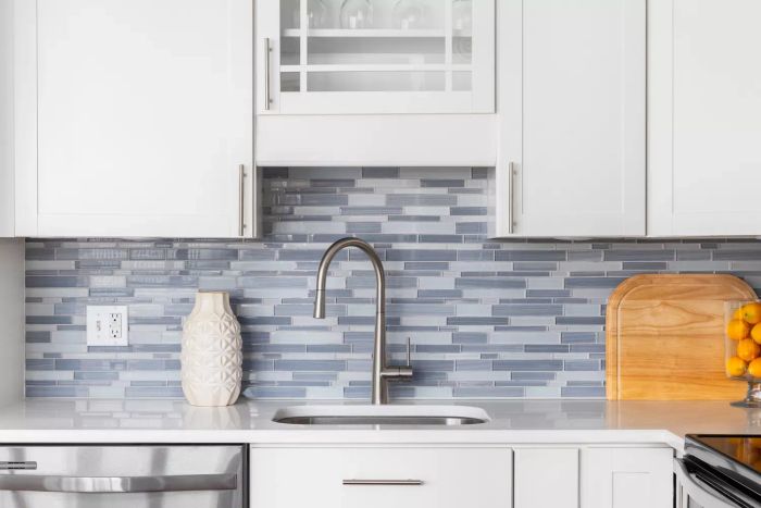 A close-up of a kitchen faucet featuring a blue glass tile backsplash, white cabinetry, decorative items on a marble countertop, and a sleek stainless steel faucet