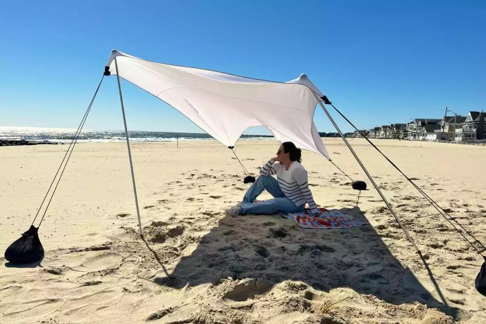 A person relaxing underneath the Neso Tents Grande Beach Tent by the shoreline.