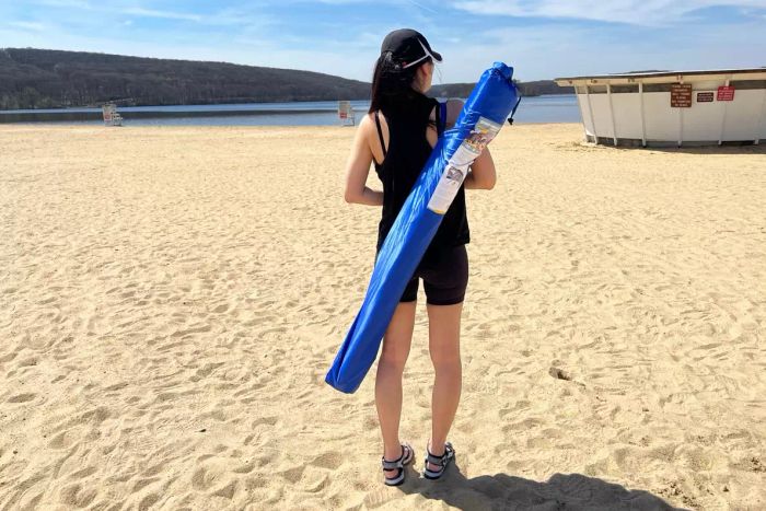 A person standing on the beach with a Sport-Brella Premiere 8-foot Outdoor Umbrella, its carrying case slung over their shoulder.