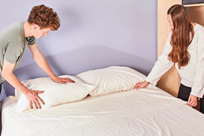 Two individuals placing The Company Store Company Cotton Rayon Made From Bamboo Sateen Sheet Set on a bed and arranging pillows.