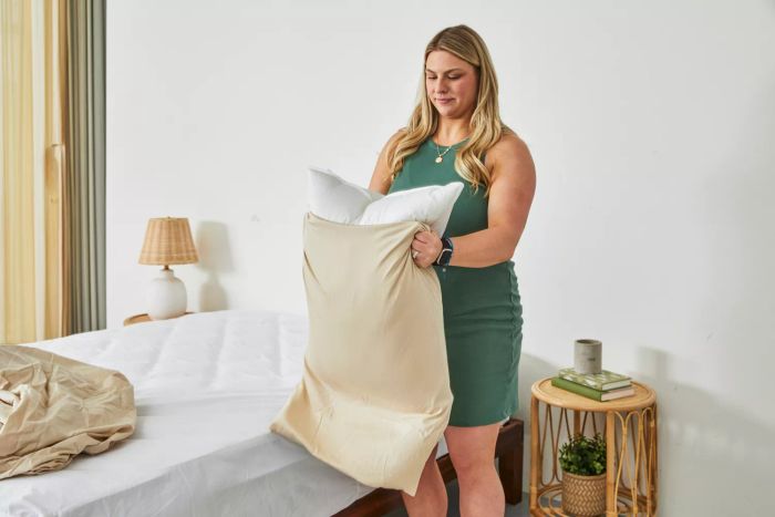 A person places a pillow inside a pillowcase made from Brooklyn Bedding Deep Pocket Bamboo Cotton Sheets, next to the bed.