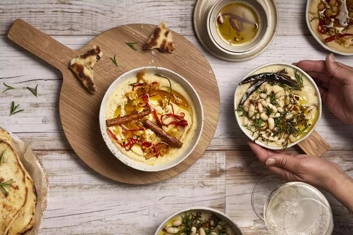 Image above displays five bowls containing chickpea hummus and cannelloni bean dip, accompanied by various spices on a wooden table with pita bread on the side. A pair of hands is seen holding one bowl, with brown being the dominant color.