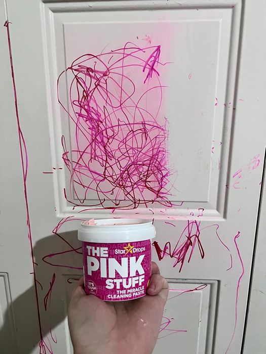 A jar of The Pink Stuff cleaning paste held in front of a door covered in pink scribbles