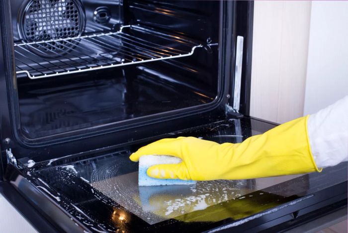 Close-up of a woman’s hand wearing yellow protective gloves, cleaning an oven door