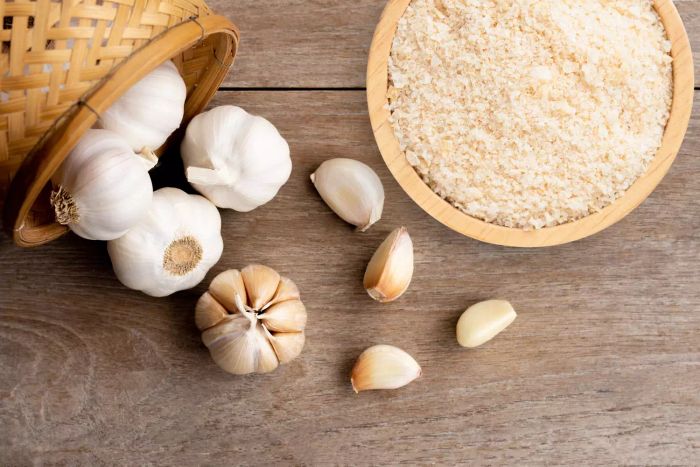 A wooden bowl filled with garlic powder alongside a bulb of garlic placed on a wooden table.