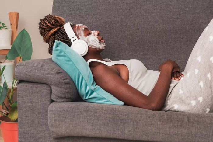 Side view of a relaxed woman wearing a beauty face mask, lying in her living room while listening to calming music through headphones