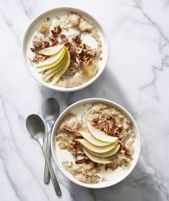 Two bowls of steel-cut oats with maple syrup, pears, and pecans, presented on a table, accompanied by two spoons.