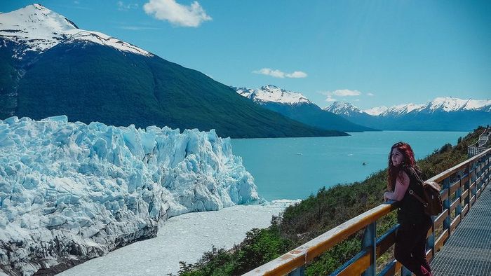 A woman standing on a bridge, gazing at a glacier surrounded by water and towering mountains