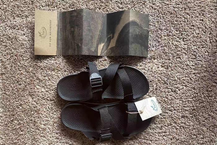 A pair of black Chaco Women’s Lowdown Slide placed beside the unfolded Chaco information sheet.