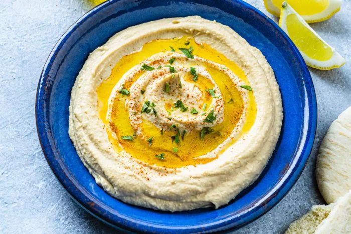 A vibrant blue plate of hummus drizzled with olive oil, surrounded by fresh veggies like celery, carrots, radish, and broccoli, with pita bread completing the plate's colorful and healthy look.