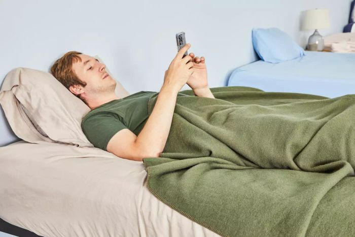 A person lying in bed, browsing on their phone, while wrapped in the Faribault Mill Pure & Simple Wool Blanket