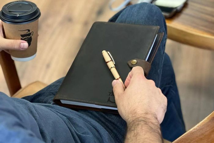 A close-up shot of a person seated with a coffee cup and a wooden pen resting on the Lightning Design Store Personalized Leather Journal.