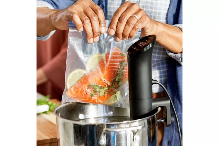 A person adding a bag of seasoned salmon, lemon slices, and herbs into a pot with the Anova Culinary Precision Cooker Nano.
