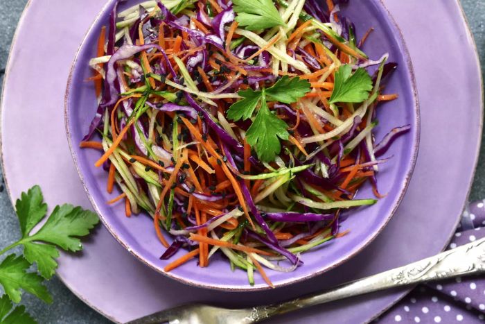 A vibrant red cabbage salad, featuring carrots and cucumbers, served in a bowl atop a dark grey slate or concrete surface