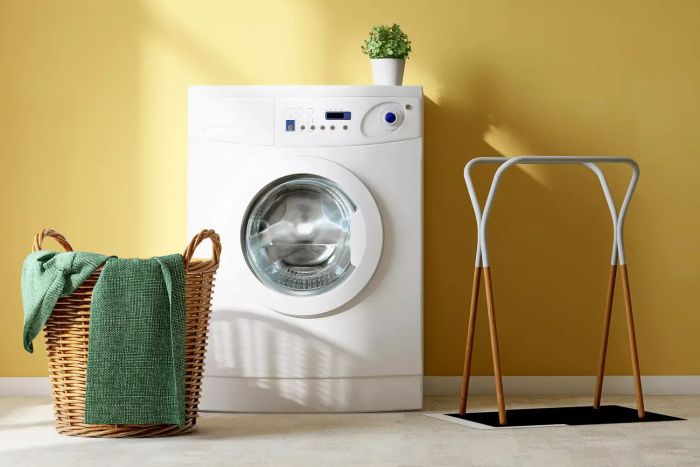 A contemporary white washing machine, a rattan basket filled with towels, and a cloth rack placed on a tiled floor in a bright yellow laundry room, illuminated by natural sunlight streaming through a window, perfect for showcasing cleaning products.