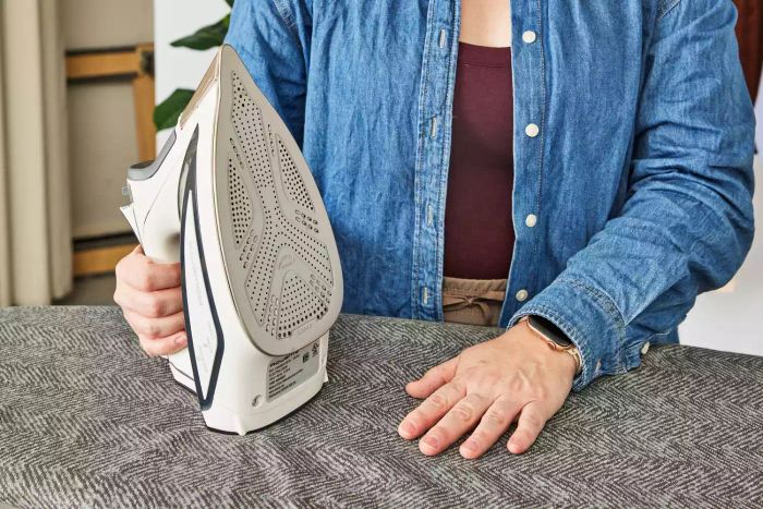 A person holding an iron in one hand while pressing down on the Bartnelli Heavy Duty Ironing Board with the other.