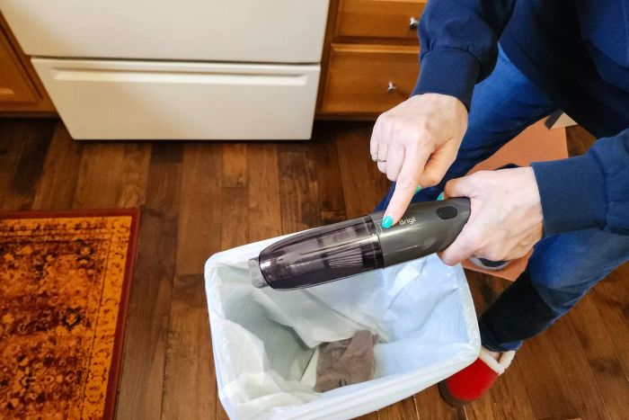 A hand pressing a button on the Brigii M5 Cordless Crevice Vacuum while holding it above a trash can
