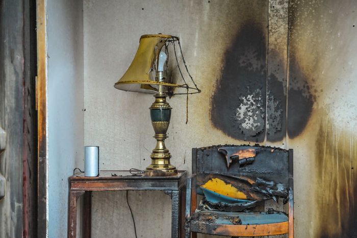 A burnt chair positioned next to a scorched lamp with a charred wall behind it