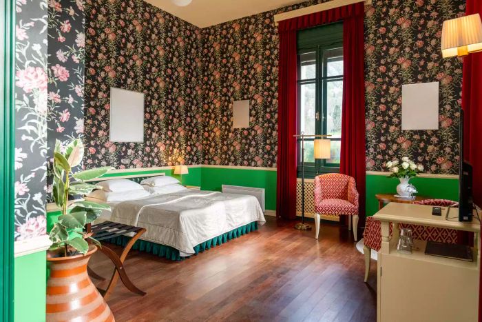 A cozy space with floral wallpaper and green wainscoting, showcasing a retro-inspired bed, elegant curtains, and timeless furniture, all glowing in the natural light streaming through the balcony door.