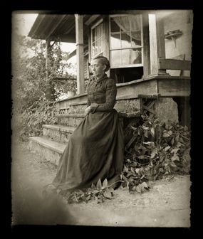 A woman dressed in Victorian-era attire, standing on the porch of her farmhouse.