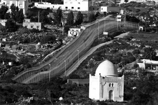 A border fence near the cemetery in Melilla, Spain, showing a large structure that symbolizes the geographic divide.