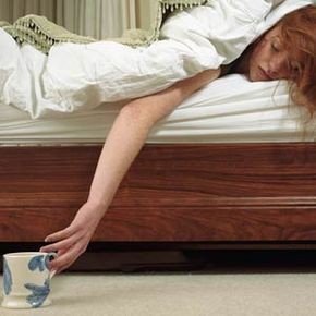 A woman, ill in bed, stretches out to grab her mug.