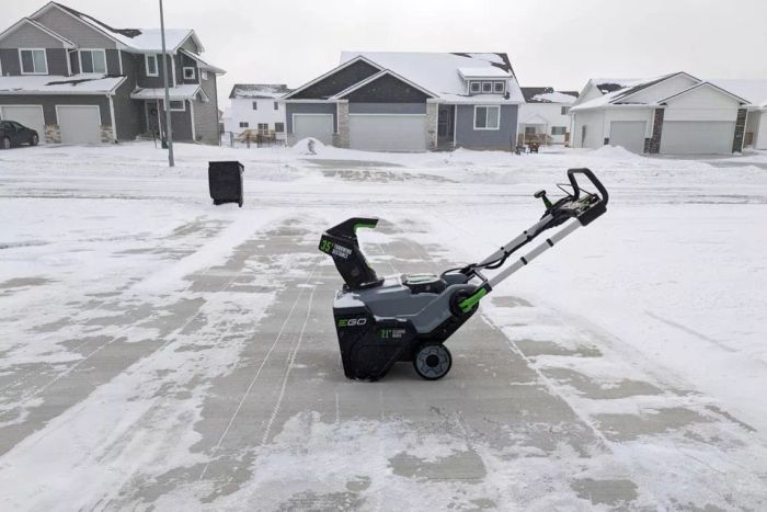 The EGO Power+ 21-Inch Cordless Single-Stage Snow Blower is shown on a driveway that has been cleared of snow.