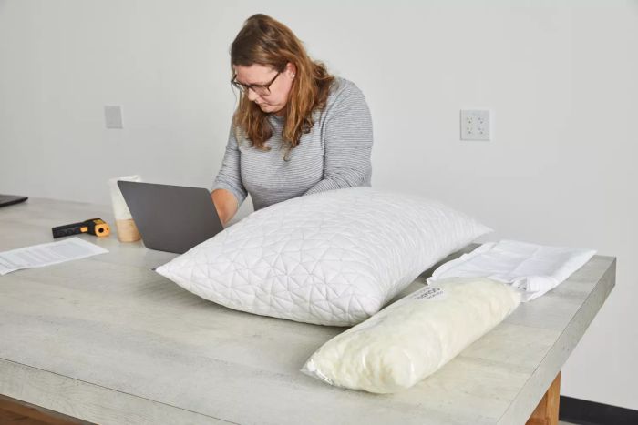 A woman typing on a laptop with a Coop Sleep Goods The Original Pillow placed on the table nearby