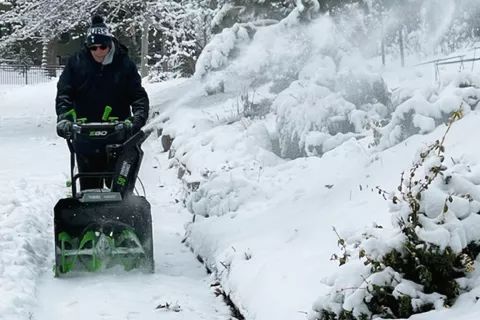 A person operates the EGO Power+ 24-Inch Cordless 2-Stage Snow Blower, clearing snow from the driveway.