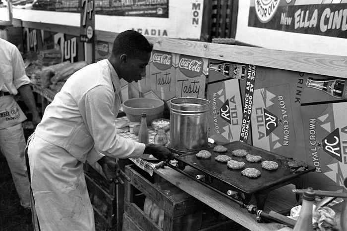 A man dressed in a full white cook’s uniform flips burger patties on a griddle at a food stand.