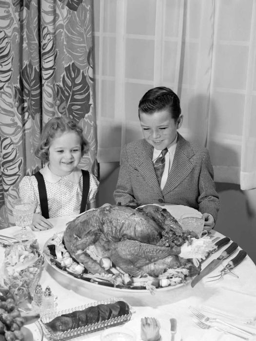 Circa 1950s: A boy and girl smile at the platter with a roast turkey on it, sitting at the dining room table. The boy is wearing a shirt.