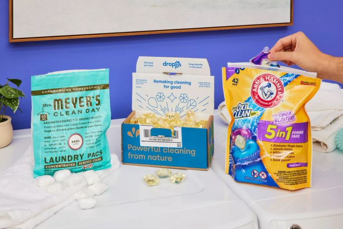 A row of laundry pod containers lined up next to a washer and dryer, with a hand reaching to grab one pod from a package