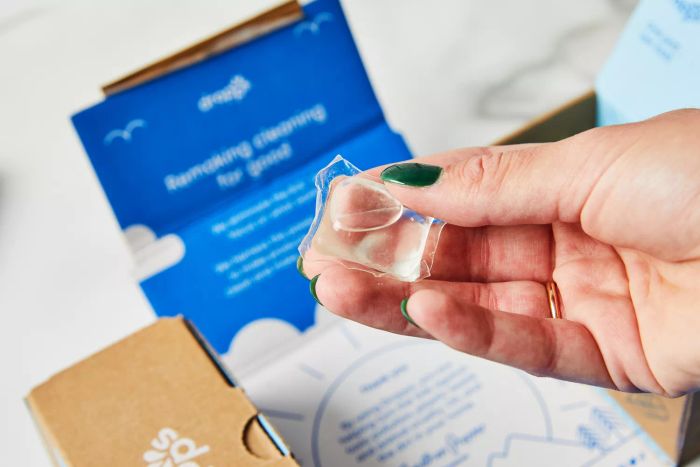 A person is holding a Dropps Sensitive Skin & Baby Laundry Detergent Pod above a box.