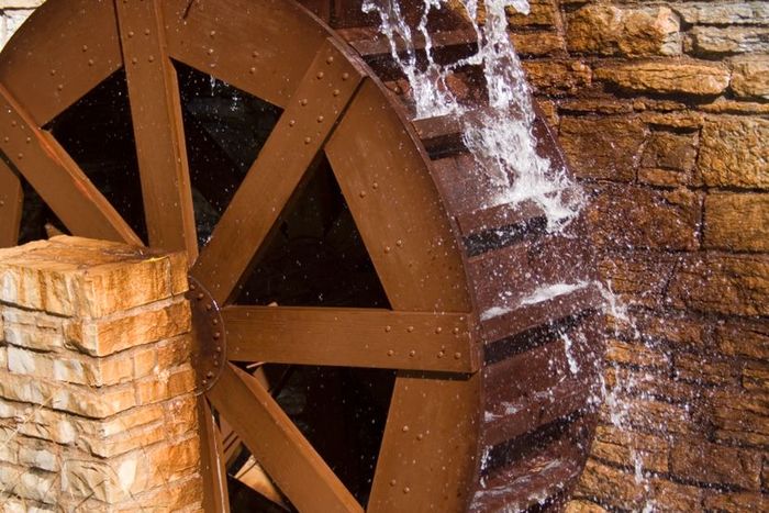 A wooden water wheel spins, mounted against the stone wall of a building's exterior.
