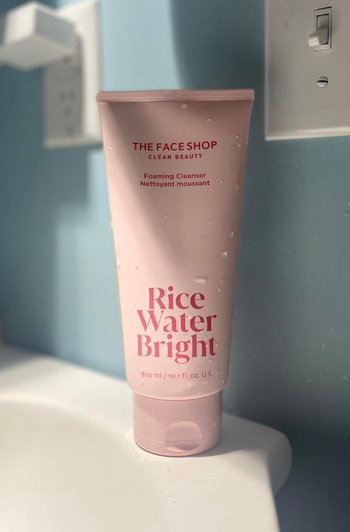 A tube of The Face Shop Clean Beauty Rice Water Bright foaming cleanser, with droplets of water on it, resting on a bathroom countertop
