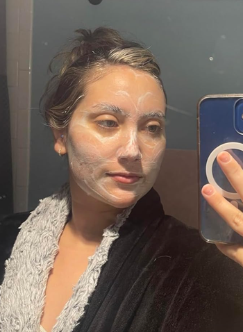 A person snapping a mirror selfie while wearing a facial mask, dressed in a comfy robe with a fluffy collar