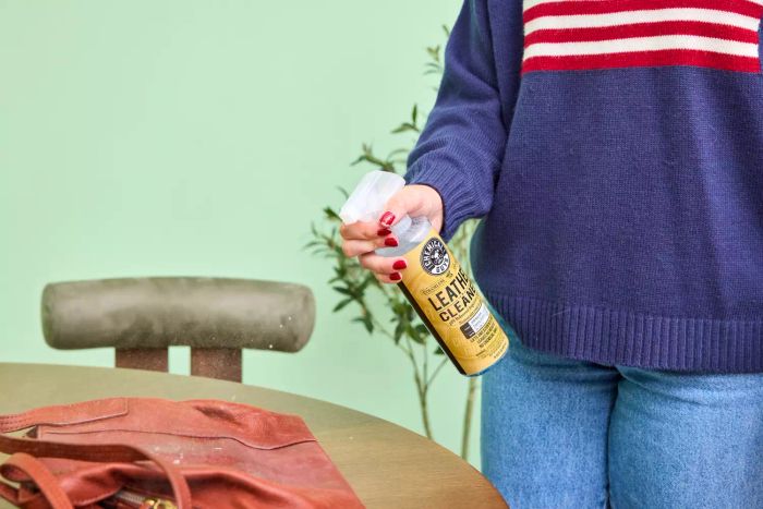 An individual sprays Chemical Guys Leather Cleaner and Conditioner Complete Leather Care onto a leather bag