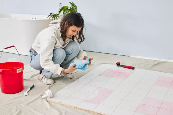 A person crouches over white tile stained with marker while testing the Rubbermaid Reveal Cordless Battery Powered Scrubber Home Kit