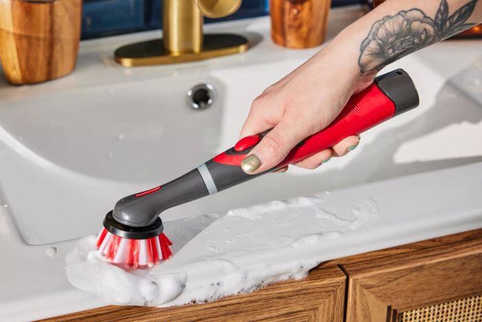 A hand holds the Rubbermaid Reveal Power Scrubber to clean a soap-covered bathroom sink