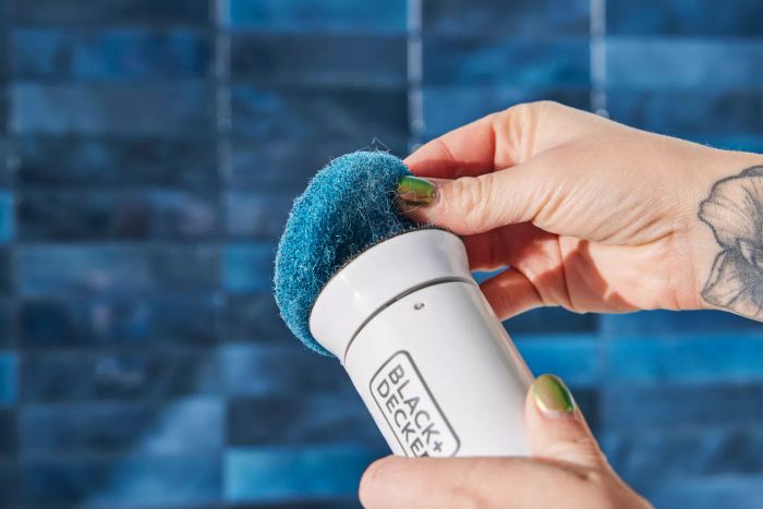 A hand detaches a scrub pad from a BLACK+DECKER Power Scrubber Brush