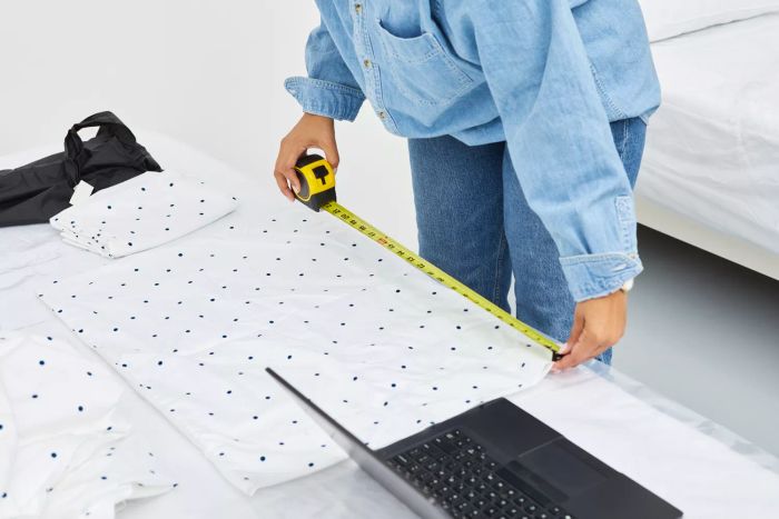 Individual measuring a pillowcase from the California Design Den Cotton Sateen Sheet Set with a tape measure, with a laptop nearby